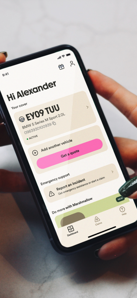 Marshmallow Insurance - Una persona che tiene uno smartphone che mostra il dashboard di Marshmallow Insurance con i dettagli della polizza auto e i pulsanti di supporto di emergenza.