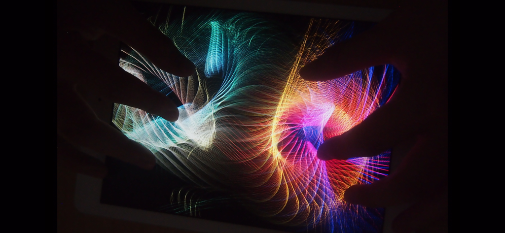 Due mani interagiscono con particelle luminose vorticose e colorate su uno schermo del tablet utilizzando il multi-touch.