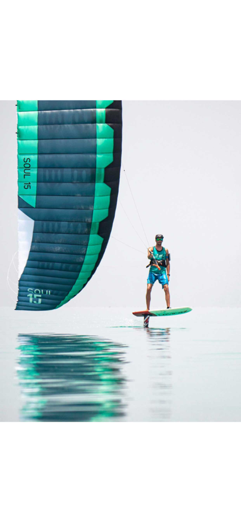 Un kitefoiler navegando en hydrofoil sobre agua en calma como un espejo con una gran cometa de foil azul verdoso y negra.
