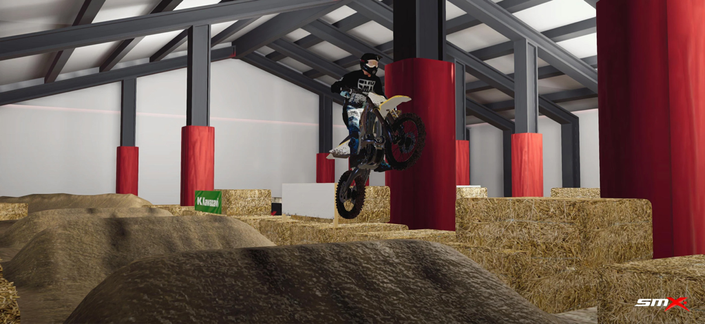 A motocross rider performing a mid-air stunt jump over dirt ramps in an indoor warehouse track.
