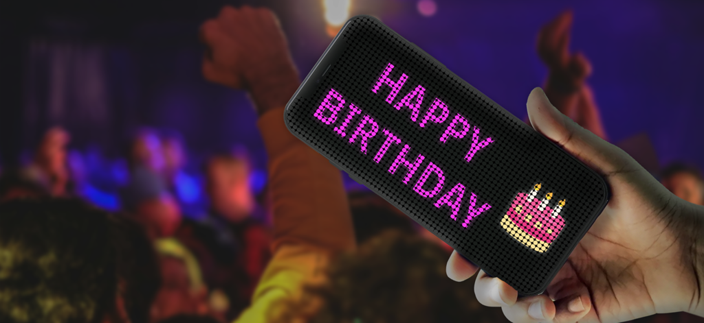 LED Banner: LED Scroller Board - Una persona sosteniendo un smartphone que muestra un cartel LED rosa desplazable con un mensaje de Feliz Cumpleaños y un emoji de pastel en un lugar abarrotado.