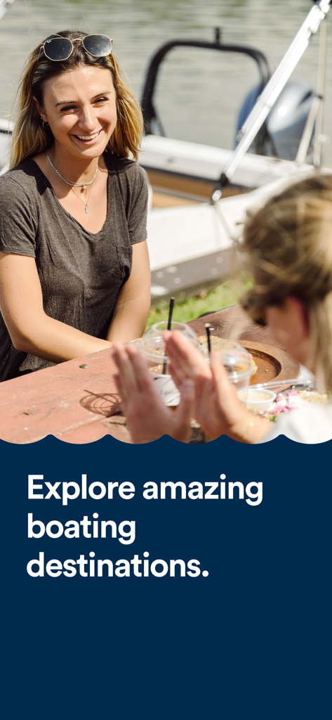 Skipperi - Una mujer sonriente disfrutando de una comida informal al aire libre en una mesa de picnic con un barco al fondo.