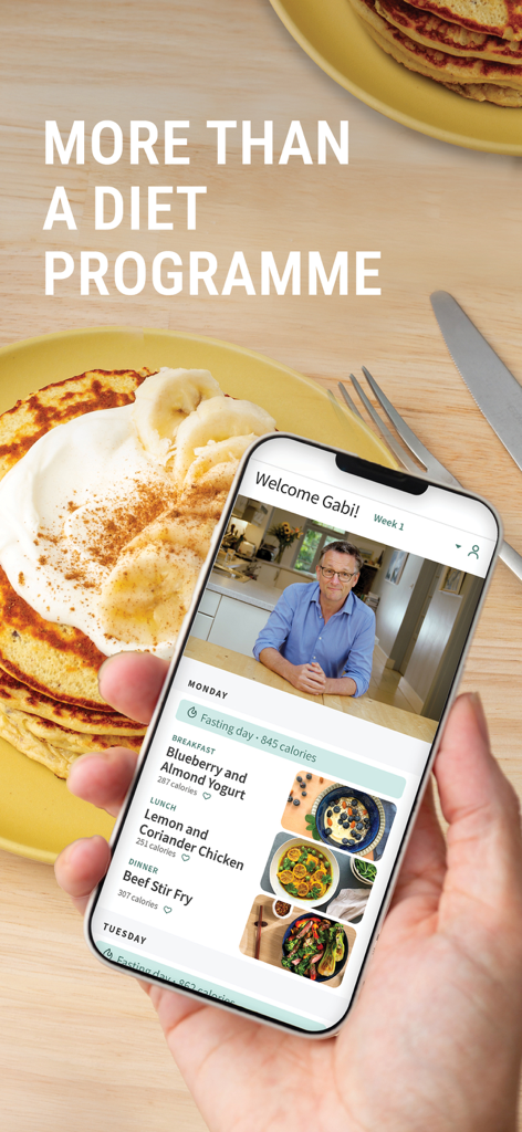 A hand holding a smartphone showing the Fast 800 app with a meal plan and a video featuring Dr Michael Mosley