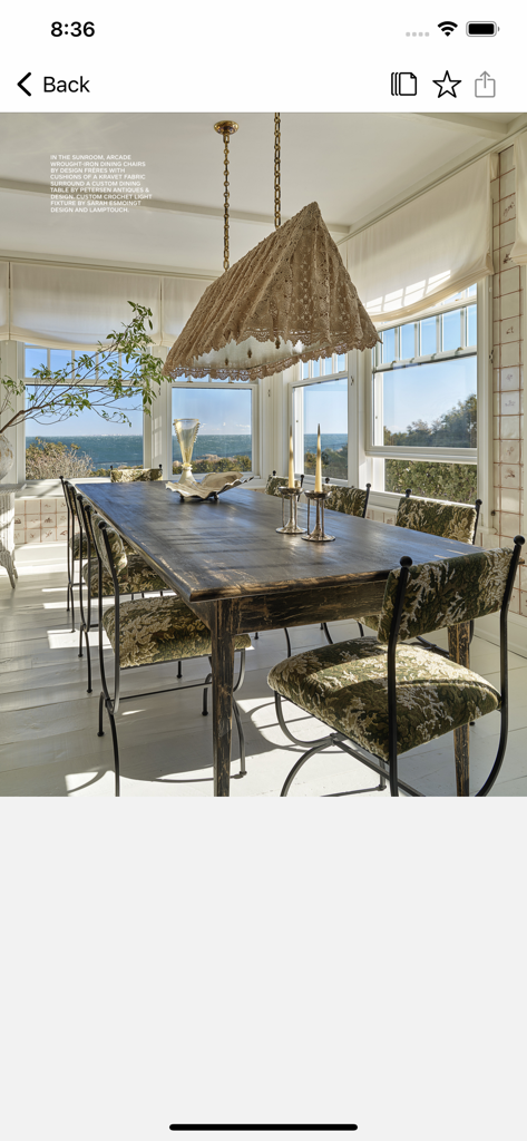 Architectural Digest - A sunroom dining area with a long wooden table, patterned chairs, and a large fringed light fixture overlooking the ocean.