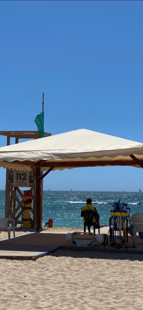 Lifeguard sitting under a white canopy at the beach with 112 SOS text visible on a wooden station