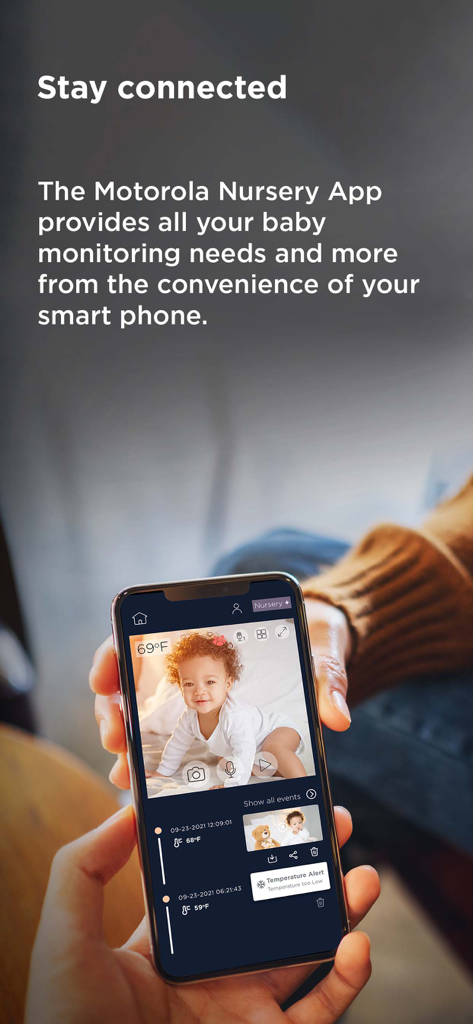 Motorola Nursery - A person holding a smartphone displaying the Motorola Nursery app with a live baby video feed and room temperature alerts