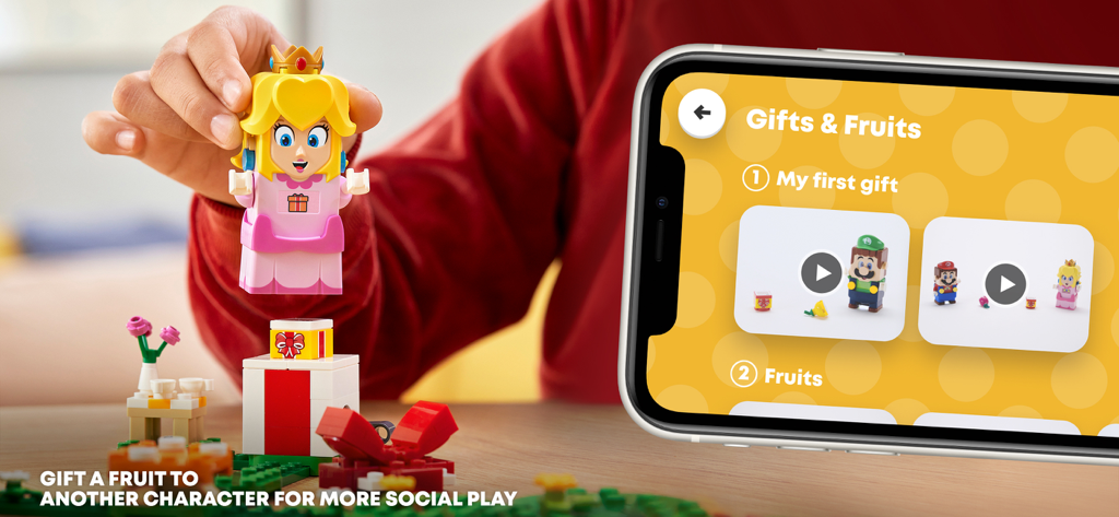A child holding a LEGO Princess Peach figure next to a smartphone displaying the LEGO Super Mario app Gifts and Fruits play screen.