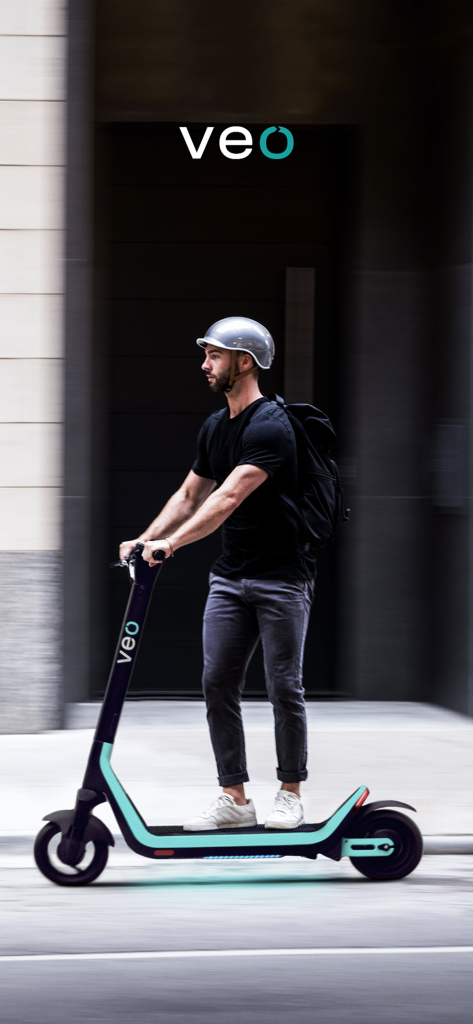A young man wearing a helmet and backpack riding a Veo electric scooter in a city