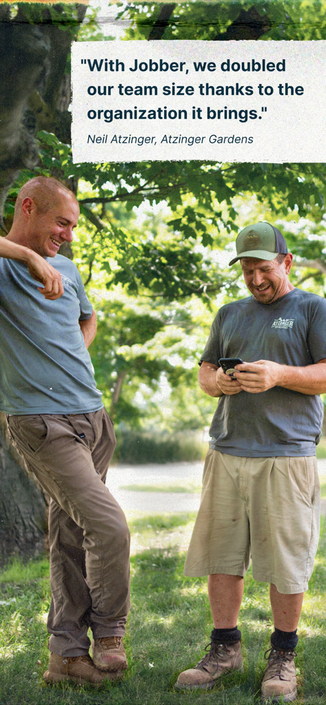 Two landscaping professionals smiling while using the Jobber app on a smartphone in a garden setting