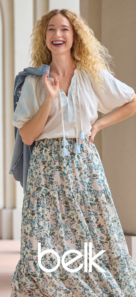 Eine lächelnde Frau, die ein weißes besticktes Oberteil und einen langen geblümten Rock aus der Belk-Modekollektion trägt.