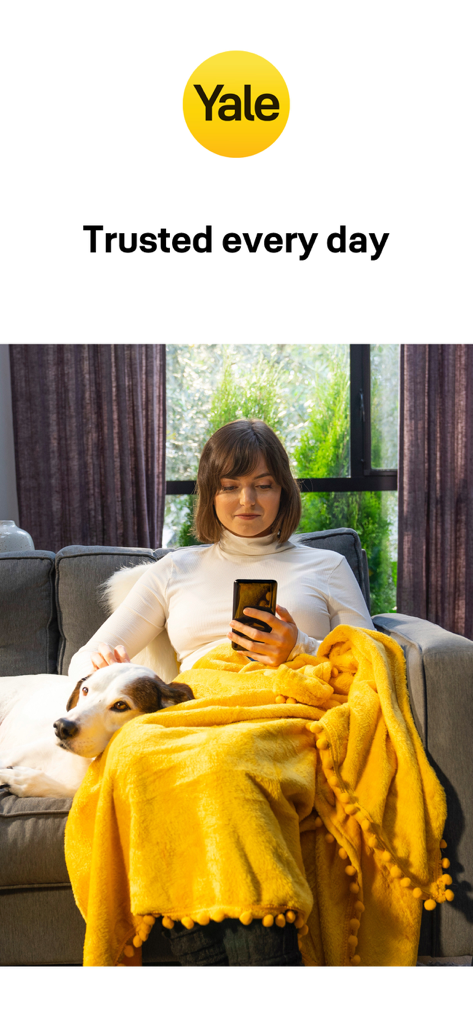 Una mujer usando la aplicación Yale Access en su smartphone mientras se relaja en casa con su perro