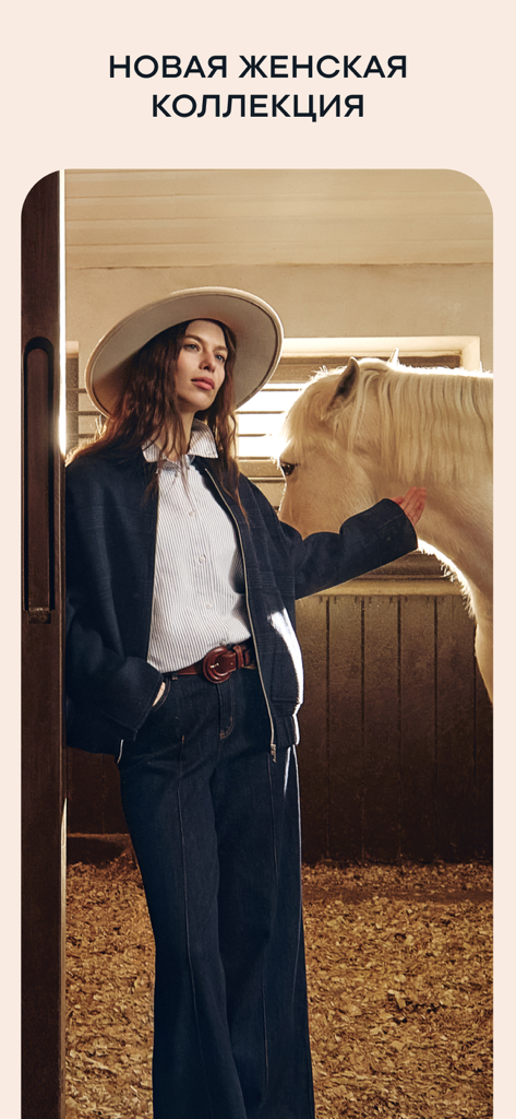 Frau in einem stilvollen Hut und Jacke posiert mit einem weißen Pferd in einem Stall für die neue BAON Kollektion.