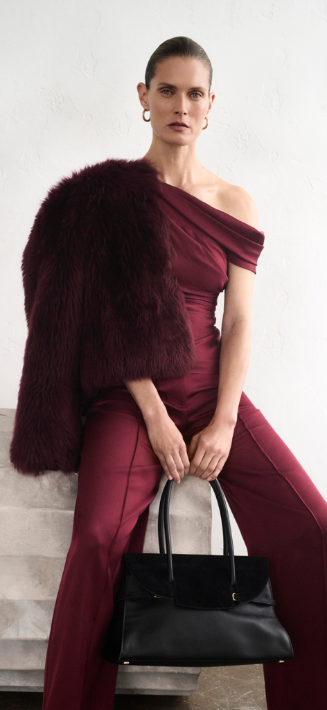 A model wearing a maroon off the shoulder outfit and fur shawl while holding a black leather handbag