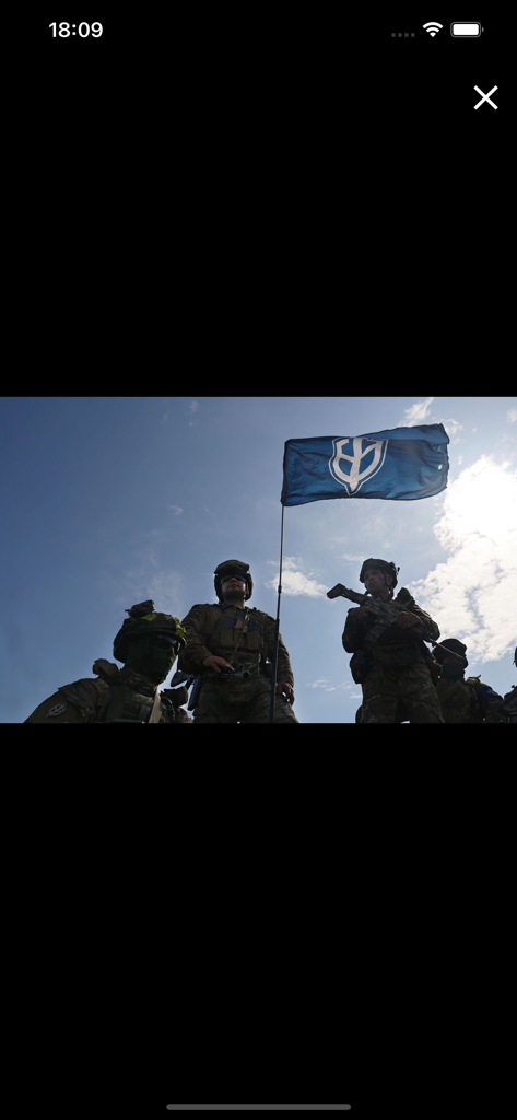 РБК Украина - новости - Soldats ukrainiens en tenue tactique tenant un drapeau bleu avec un emblème blanc sous un ciel bleu