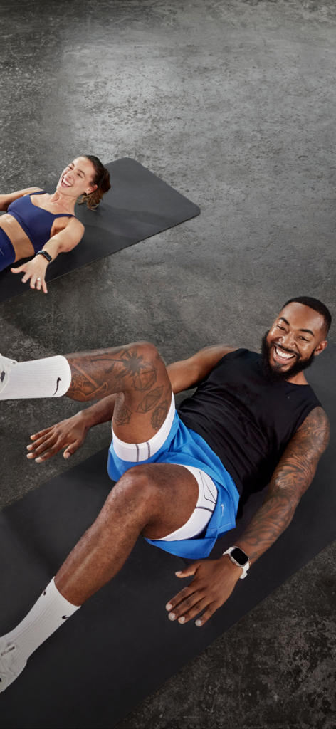 A man and a woman smiling while performing core exercises on yoga mats during a Nike Training Club session.