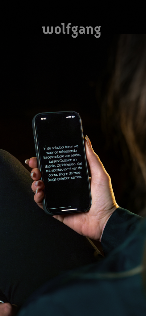 Wolfgang - 暗いコンサートホールでライブクラシック音楽の字幕を表示するWolfgangアプリを搭載したスマートフォンを持っている人