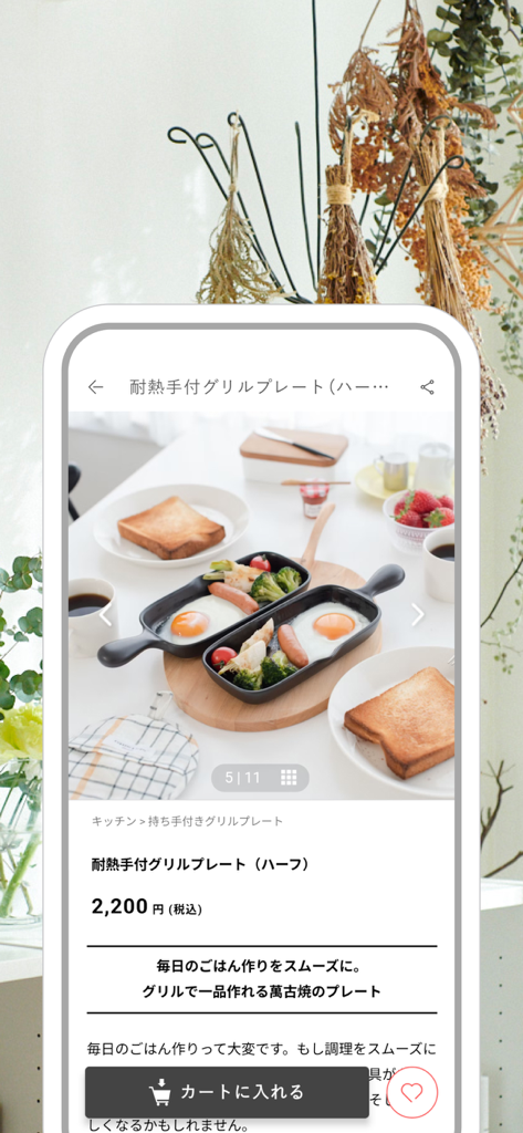 Une page produit dans l'application de style de vie nordique montrant une plaque de cuisson en céramique utilisée pour un petit-déjeuner d'œufs et de saucisses sur une planche en bois.