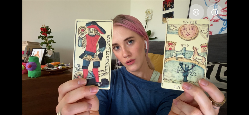 A woman with pink hair holding up two tarot cards during a video date