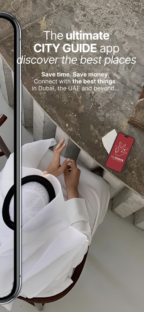 Una persona con vestimenta tradicional emiratí en una mesa de piedra con un smartphone mostrando el icono de la aplicación The HUNTR.