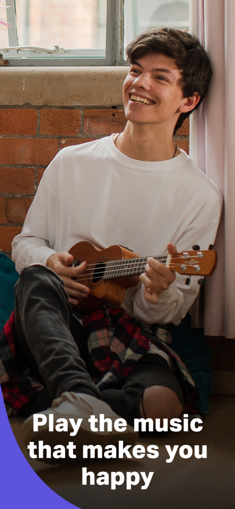 Un joven sonriente tocando un ukelele sentado contra una pared de ladrillos.