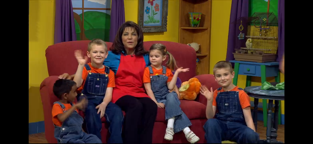 X IPTV Player Pro - Eine Frau und Kinder winken, während sie auf einer roten Couch während einer Kindersendung sitzen