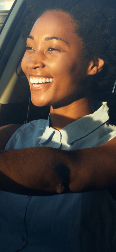Evopay Africa - 太陽が沈む時間帯に車を運転しながら笑顔を見せる、アフロヘアの幸せそうな若い女性