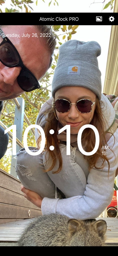 Atomic Clock PRO - Digital clock display with a custom personal photo background featuring a couple and a quokka in the Atomic Clock PRO app.