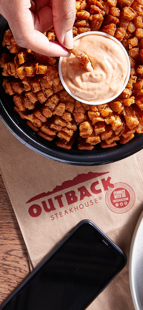 Outback Steakhouse - Mano mojando una Bloomin Onion en salsa junto a un smartphone y una bolsa de Outback Steakhouse