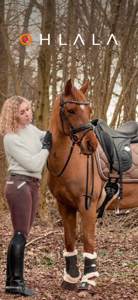 Ohlala Sellerie - Une cavalière en tenue équestre se tient à côté d'un cheval bai sellé dans une forêt avec le logo Ohlala.