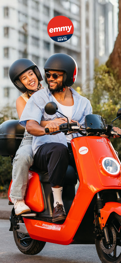 Ein lächelndes Paar mit Helm fährt auf einem roten emmy Elektroroller durch die Stadt.