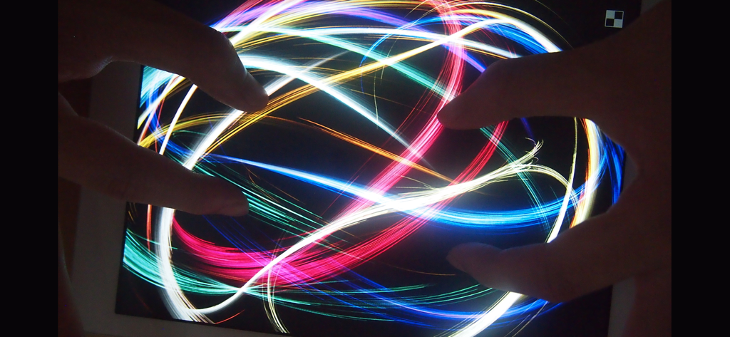 Una persona usa più dita su un tablet per creare vibranti scie luminose al neon nell'app Atomus