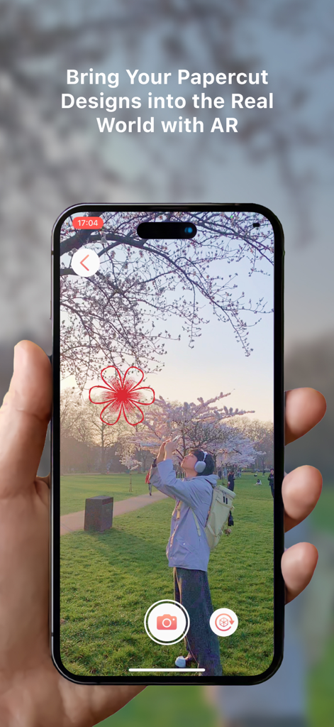 Paper Bloom - A smartphone screen showing the Paper Bloom app using AR to place a red papercut flower design into a real world park setting with cherry blossoms.