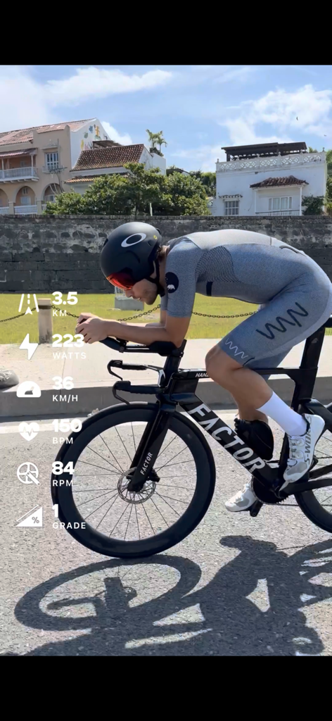 A cyclist riding a triathlon bike with live performance data telemetry overlays for speed, power, and heart rate