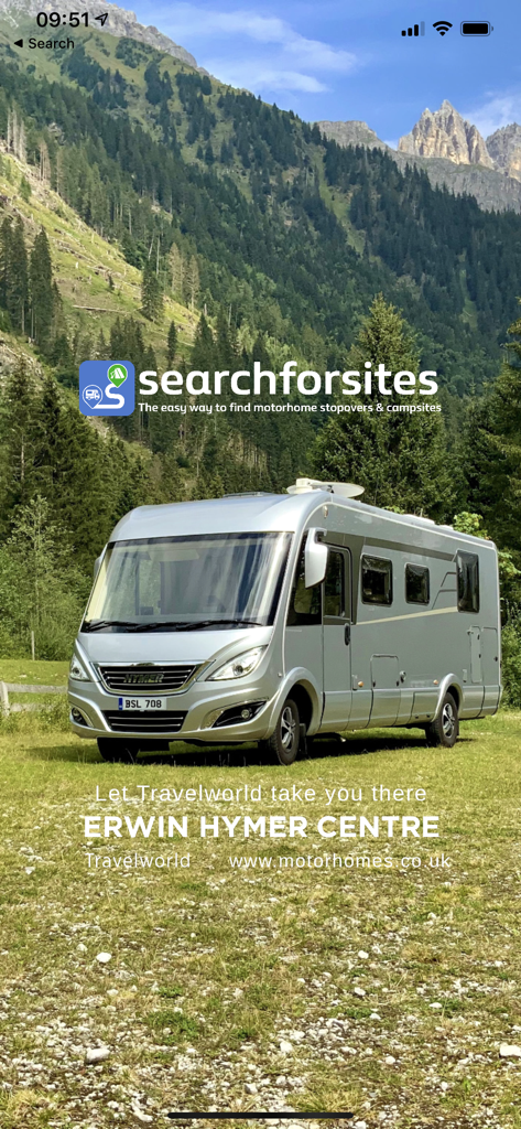 A silver motorhome parked in a grassy field with a backdrop of mountains and forests featuring the searchforsites logo.