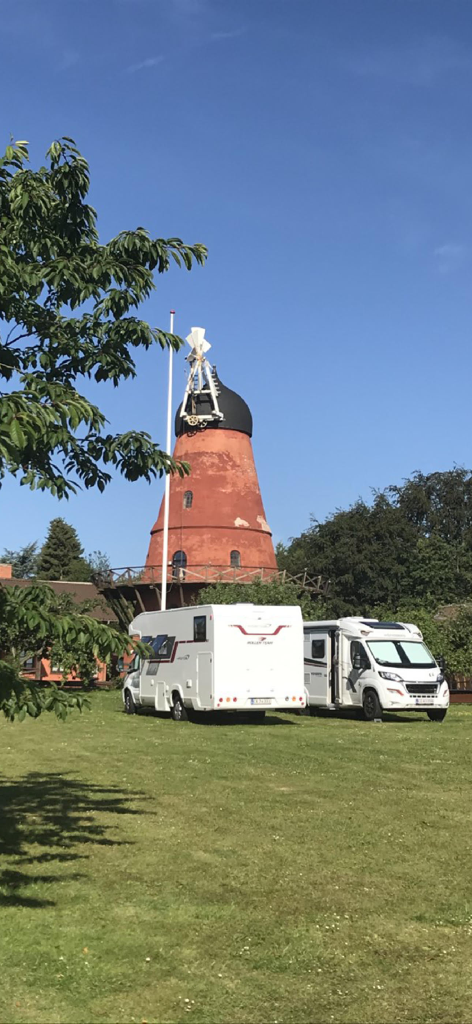 Pintrip - motorhome stopovers - Two white motorhomes parked on a green field in front of a tall, historic red brick windmill in Denmark under a clear blue sky.
