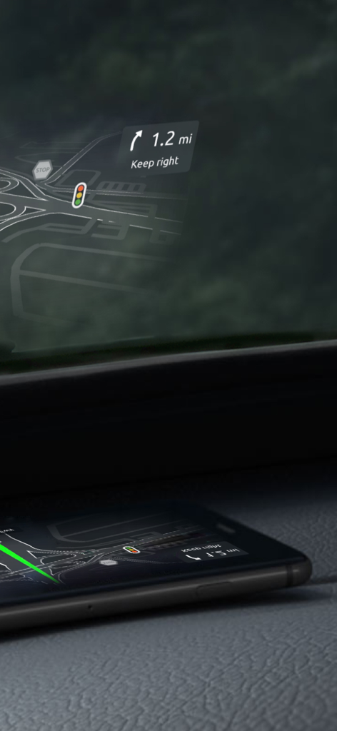 A smartphone on a car dashboard reflecting a navigation heads-up display onto the windshield showing road directions and traffic signs.