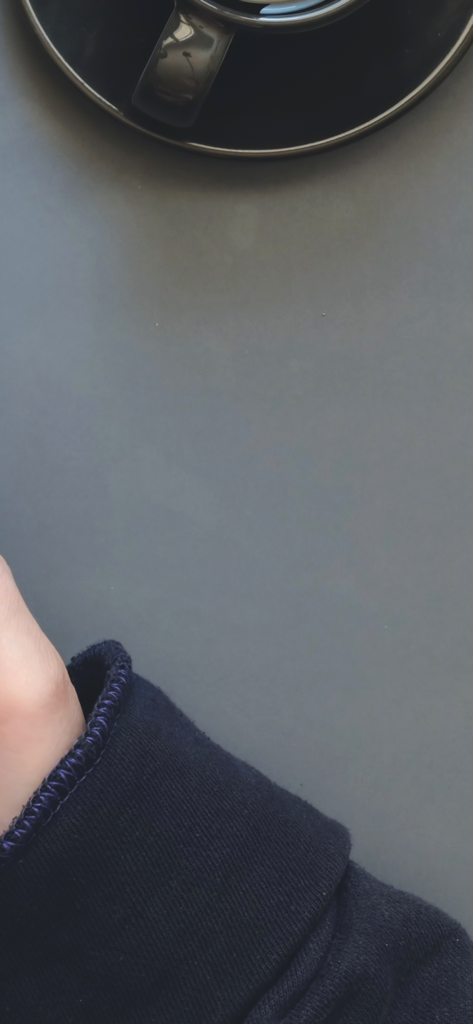 A minimalist overhead view of a coffee cup on a dark desk next to a person's arm in a sweater, symbolizing a focused work environment