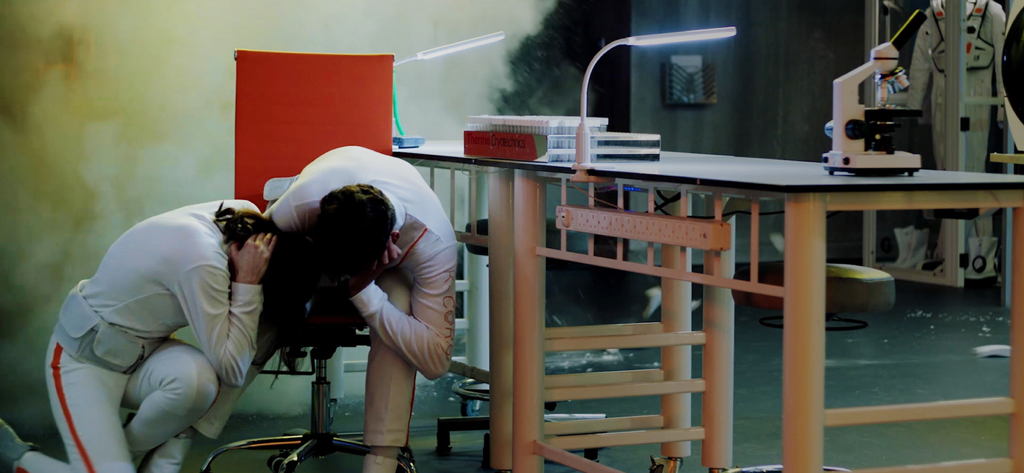 Dos personajes con trajes blancos agachados en un laboratorio de alta tecnología lleno de humo.